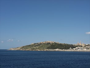 The coastline and hills with buildings in the background and a clear blue sky, the island of Gozo