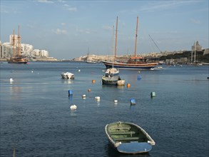 Several boats and a large sailing boat on the calm sea in front of a historic town and colourful