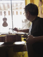 A luthier uses tools on a violoncello near a bright workshop window, surrounded by crafting