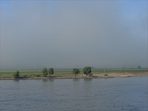 A calm river stretches along a foggy bank with green fields and trees, fog at the rhine river with