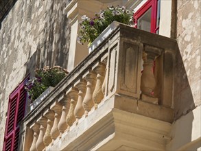 A small balcony with flowers and shutters adorns an old stone wall, the town of mdina on the island