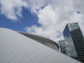 Arched white structure and glass facades of skyscrapers under a blue, partly cloudy sky, modern