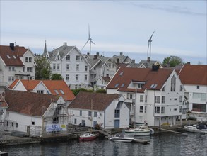 White houses with red roofs stand on the shore of a coastal town. Some wind turbines can be seen in