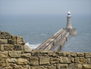 Stone wall and a lighthouse on a long pier jutting into the sea, under morning mist and blue sky,