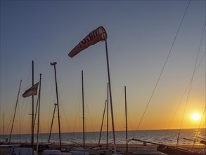 Sunset at the beach harbour with several sailing boats and waving flags, calm sea and clear sky,
