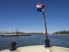 View from a boat on the water with a waving flag, sailing boats in the harbour and a bright blue