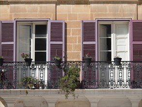 A mediterranean balcony with purple shutters and potted plants in front of two open windows, the