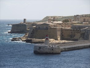Stony coastline with historic lighthouse and old fortifications on blue water, Valetta, Malta,