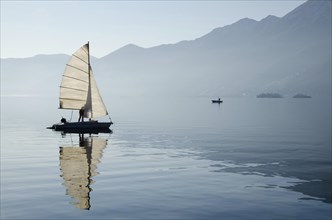 Old Sailing Boat Reflected on Lake Maggiore with Brissago Islands and Mountains in a Misty Sunny