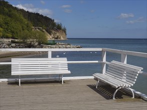 White benches on a jetty, behind them the sea and a wooded cliff, spring on the Baltic coast with