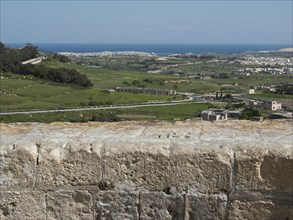View over a stone wall to fields and the sea under a blue sky, the village of mdina on the island