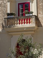The balcony of an old building with red windows and colourful flower pots, the town of mdina on the