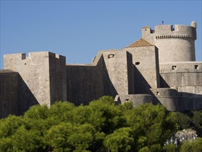 An imposing medieval fortress with high walls and towers rises into the blue sky, surrounded by
