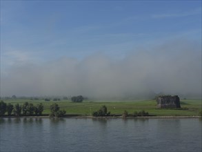 A ruin lies next to a river, surrounded by green fields and fog, ships on the rhine river in the
