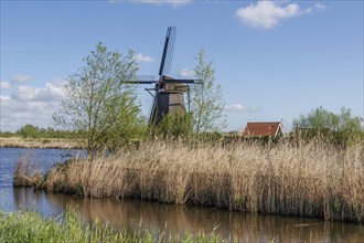 Windmill in a natural setting with river, reeds and a house, under a blue sky, windmills of