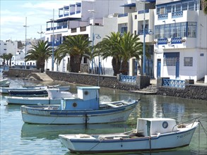 Several boats float in the quiet harbour, surrounded by white buildings with blue accents and palm