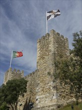 Medieval castle with Portuguese flag on a watchtower spire, surrounded by trees under a clear sky,