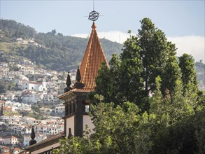 A tower with a pointed top towers over trees and houses on a hill, Madeira, Portugal, Europe