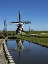 Large windmill with its reflection in a pond, surrounded by people, under a clear blue sky,