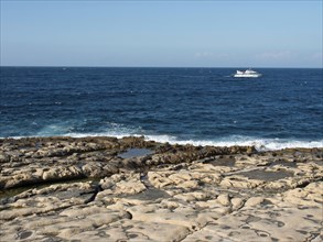 Rocky coastline with crashing waves and a boat in the distance under a clear sky, Valetta, Malta,