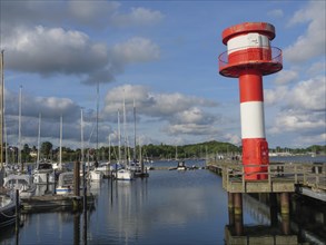 Tall red and white lighthouse at the edge of a harbour with calm water, surrounded by sailing