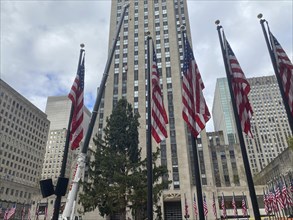 Several american flags waving in front of a skyscraper and other buildings, sky slightly cloudy,