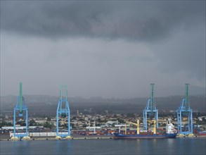 Large blue harbour cranes lifting containers, cloudy sky and a ship in the harbour industrial area