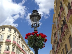 Wrought iron street lamp with red flowers against a clear blue sky and surrounding buildings, the