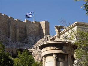 An ancient castle with a waving Greek flag and a decorative column in the foreground under a clear