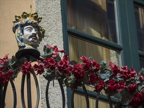 Ceramic bust on a balcony with red flowers in front of a window on a rustic facade, The old town of