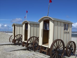 Beige changing cabins with wooden wheels stand on a sandy beach under a blue sky, historic bathing