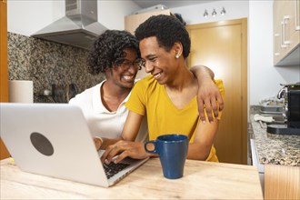Gay couple laughing and embracing while using laptop drinking coffee sitting in the kitchen at home
