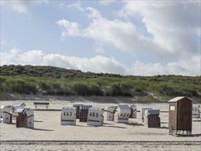 Beach chairs are scattered on an empty beach with green dunes and cloudy skies, Spiekeroog,