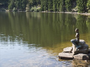 Statue of a seated figure looking at the water surface of a calm lake, small forest lake in the