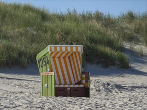 A single green beach chair with yellow and white stripes stands on the beach in front of a grassy