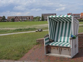A green and white beach chair stands on a paved square with houses in the background, Baltrum
