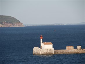 White lighthouse with red roof stands on a pier in the calm blue sea under a clear sky, la seyne