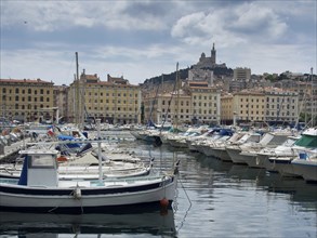 Harbour with many boats, behind it colourful buildings and a hill with a church, Marseille at the