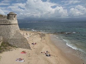 People relaxing on a sandy beach next to a fortress with sea and cloudy sky, Corsica, Ajaccio,