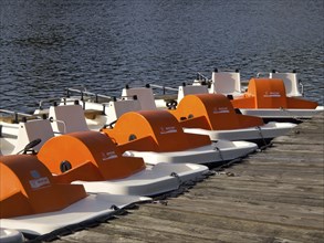 Row of orange-coloured pedal boats along a wooden jetty at the lake, small lake with hiking trails