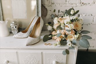Elegant wedding setup with glittery shoes, floral bouquet, and rings on a rustic white dresser
