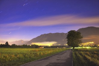 Beautiful Road with a Tree and Mountain at Night in Long Exposure with Star Trails and Clouds in