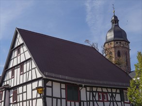 A half-timbered house with red roof tiles next to a gothic church with a tower under a blue sky,