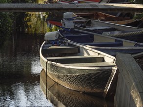 A boat lies under a wooden bridge in a shady area of water, surrounded by dense vegetation, boats