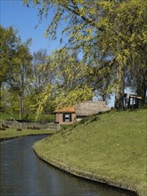 A small cottage stands on a riverbank, surrounded by green grass and flowering trees under a blue