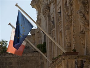 Two flags, one of the EU and one of Malta, in front of historic stone architecture, the town of