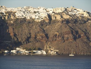 A cluster of white buildings on a cliff dramatically jutting out over the calm sea, brown volcanic