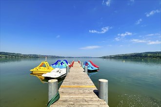 A jetty leads to several colourful pedal boats on a calm lake under a clear summer sky, Sempach,