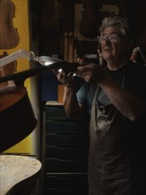 In a dark workshop, a luthier examines a violin under the light of a desk lamp, surrounded by tools