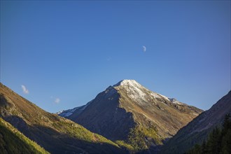 A snow-covered mountain peak under a blue sky with a small crescent moon, surrounded by mountains